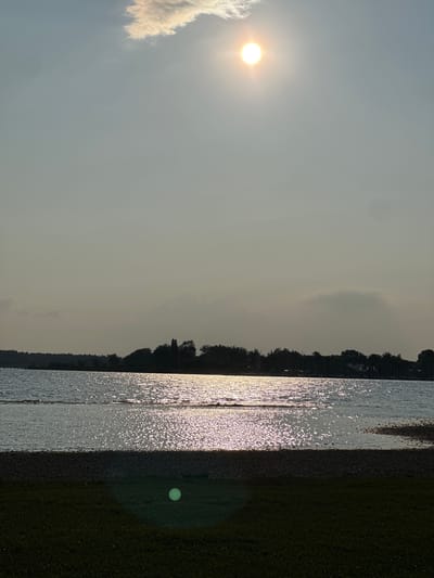 This is a vertical, eye-level shot of a serene lakeside scene in Chieming, Germany, taken in the late afternoon or early evening. The sun, positioned high in the sky, casts a bright, warm glow, creating a shimmering reflection on the water's surface. A solitary cloud drifts near the sun, tinged with a pale yellow hue.
The foreground features a patch of dark green grass, with a gravelly shore bordering the lake. The water itself is calm, with gentle ripples reflecting the sunlight. Across the lake, a dense line of trees forms a dark silhouette against the horizon, with a few structures, possibly buildings or boats, visible amidst the foliage on the right side. The overall atmosphere is peaceful and tranquil, highlighting the natural beauty of the Chieming area. There are no people or animals present in the frame.