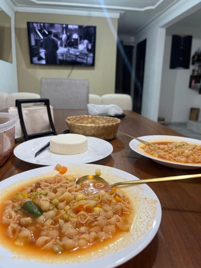 Two bowls of pasta soup sit on a wooden table, one partially obscured. A gold ladle rests in the closer bowl. A small white plate holds a cylinder of white food, possibly cheese, alongside a knife. A woven basket is visible. The background features a TV on a wall, a cream-colored sofa, and a gray chair.