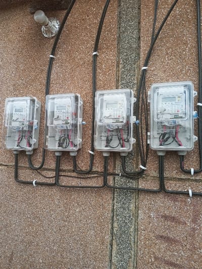 Four "HEXING" brand digital electricity meters are horizontally mounted on an exterior building wall in Konongo, Ghana. Each meter is enclosed within a clear plastic housing, displaying an LCD screen and a keypad on its face, with internal wiring visible. Black electrical conduit cables are neatly routed along the granular, light brown textured wall, secured by numerous white plastic clips. These cables connect to the top of each meter, extending downwards to the bottom, and then continue horizontally along the wall. A distinct vertical strip of darker, greyish, rough-textured material runs through the wall's center, serving as a structural element or joint. An exposed incandescent light bulb in a metallic fixture is affixed to the wall above the leftmost meter. The entire assembly indicates an organized utility installation.