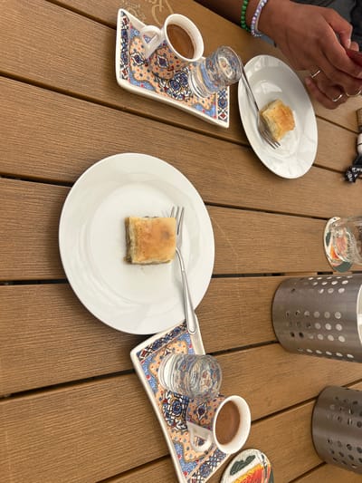 This medium close-up photograph captures an outdoor table setting, likely at a cafe or restaurant in Liberec, Czechia. The scene features three servings of baklava and Turkish coffee on a light brown, slatted wooden table.
In the foreground and center, a square piece of baklava is presented on a white oval plate with a silver fork. To the left, a person's hands are visible, holding a red smartphone. This individual wears several rings and two beaded bracelets (one in white and purple, another in green). Adjacent to their hands is one of three traditional Turkish coffee sets, comprising a small patterned cup filled with dark coffee and a tiny glass of water, all placed on an ornate, rectangular tray with colorful geometric patterns. A similar coffee set and another baklava plate are arranged on the right side of the table.
Two empty, clear drinking glasses are visible, one in the upper middle and another beside the right coffee set. In the background, two cylindrical silver utensil holders with perforated sides stand on the table. A small, circular coaster with an abstract design is partially visible on the far right. A small black item near the top center shows partially obscured white text, reading "t.we" and what might be the beginning of another word like 'b' or 'a'.
The activity depicted is a casual break or social gathering, with individuals enjoying dessert and coffee. One person is engaged with their mobile phone. The lighting suggests daytime, with soft illumination indicative of an overcast sky or shaded outdoor area. The overall ambiance appears relaxed and pleasant, suited for outdoor dining.