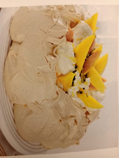 The image displays a close-up, overhead view of a meringue-based dessert, likely a pavlova, arranged on a white plate. The meringue base is a pale, golden-brown color with a characteristic cracked surface. It is generously filled and topped with whipped cream and bright yellow fruit slices, possibly mango or passionfruit, along with dark seeds, suggesting passionfruit. The dessert is artfully presented with the fruit arranged in a circular pattern. The background is a plain white surface. The image does not depict any elements that would allow for its location to be identified within Rumaithiya, Kuwait, or any other specific geographical location.