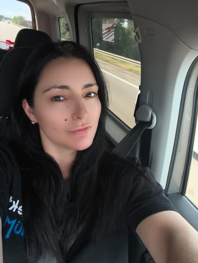 The image shows a woman with long, dark hair, wearing a black t-shirt, sitting in the passenger seat of a car. She is looking at the camera, and her face is slightly tilted. The interior of the car is visible, including a seatbelt, a car door, and windows. Outside the car, a road and trees can be seen. The overall impression is that of a candid selfie taken during a car journey. There is no visual information in the image that can be used to infer the location as Mengen, Germany.