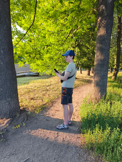 A young male wearing a light-colored t-shirt, dark shorts with a camouflage pattern, a blue baseball cap, and sandals stands on a dirt path in a park. He is holding a smartphone in his hands, looking down at it. A black strap bag is slung across his chest. The scene is set in a park with large trees, lush green foliage, and grass. The sun is casting long shadows, indicating it is late afternoon or early evening. The weather appears to be warm and sunny. In the background, a light-colored fence and some concrete structures are visible. The overall mood is relaxed and casual. The word "UNMATCHABLE" and possibly "LIMITS" are visible on his shorts.