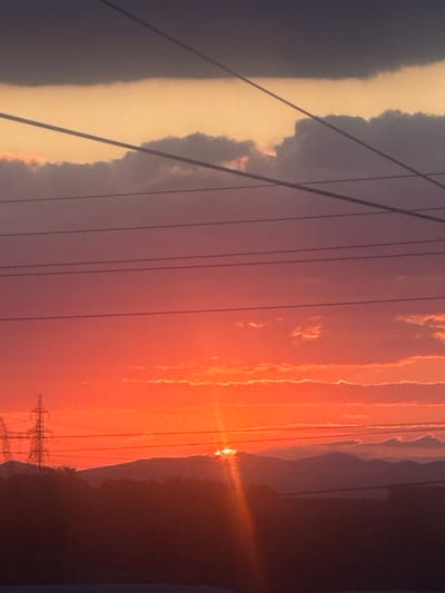 The image captures a dramatic sunset over Miriantsi, Bulgaria, with the sun partially obscured by a mountain range in the distance. The sky is ablaze with vibrant oranges and reds, transitioning to shades of pink and purple as it reaches higher. Wispy clouds are scattered across the sky, catching the last rays of sunlight. Several power lines are visible in the foreground, stretching horizontally across the frame. In the middle ground, the silhouettes of trees and hills are visible, with two power line towers standing out against the vibrant sky. The overall impression is one of natural beauty and the fading light of day.