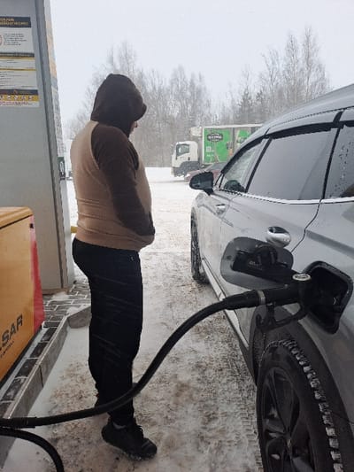 A person in a brown hooded jacket and black pants fills a gray car with gas at a snowy station. The gas pump nozzle is inserted into the car's fuel tank. Snow covers the ground, and trees and vehicles are visible in the background. The scene appears to be cold and overcast.