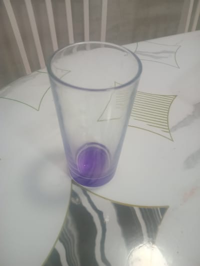 A clear glass cup with a purple liquid at the bottom lies on a white surface. The surface has a decorative pattern with green lines and a marbled effect. In the background, a grey striped pattern is visible. The lighting suggests it is daytime. The scene appears to be indoors, likely on a table. No people are visible, and no text is present.