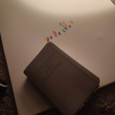 The image shows a closed, grey hardcover book with the title "NEW WORLD TRANSLATION OF THE HOLY SCRIPTURES" on the front cover. The book is lying on a white canvas, with a few strokes of colored pencil visible on the canvas surface. The location, based on the coordinates provided, is Happy Valley, Oregon, United States. The image is lit from the top right, casting shadows across the book and the canvas. The scene is a simple still life, capturing a moment of quiet contemplation or artistic creation.