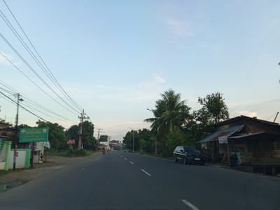 The image depicts a roadside scene in Gampongrayadagang, Indonesia, under a pale blue sky with some wispy clouds. A paved road, marked with white dashed lines, runs through the center of the frame, curving slightly to the right. On the left side of the road, there is a building with green and white accents, a large green sign, and power lines overhead. Beyond the building, there is a dense line of trees. On the right side of the road, there is a rustic wooden structure with a corrugated metal roof, possibly a shop or dwelling, and a dark colored SUV parked in front of it. Further down the road, more trees and some buildings are visible, and several motorcycles are seen on the road, indicating moderate traffic. The overall impression is of a rural or semi-urban setting in Indonesia during the daytime, likely late afternoon or early morning given the soft lighting.