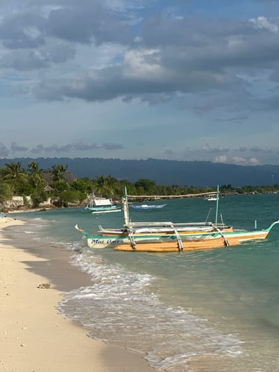 An over-the-shoulder shot shows a beach scene in Saavedra, Philippines. A tan sand beach is in the foreground, with waves gently lapping the shore. A narrow rock protrudes from the sand near the water's edge. A small, outrigger boat named "Mae Anon" is tied to the shore, bobbing in the turquoise water. The boat is painted with orange, green, and white stripes. Beyond the boat, several other boats are visible on the water, some with white awnings. In the distance, lush green trees and palm trees line the shore, with thatched-roof buildings nestled amongst them. A chain of dark, misty mountains forms the horizon, silhouetted against a sky filled with dramatic, grey clouds.