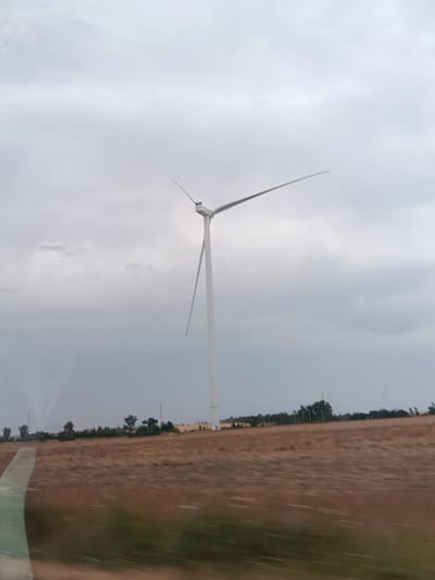 A solitary white wind turbine stands prominently in a field of dry, brown grass under a cloudy sky. The turbine's blades are angled upwards and to the right, partially obscuring the sky. In the background, a line of trees and other, smaller wind turbines are visible along the horizon. The foreground shows a blurred motion effect, suggesting the image was taken from a moving vehicle. The scene appears to be in Sychavka, Ukraine.