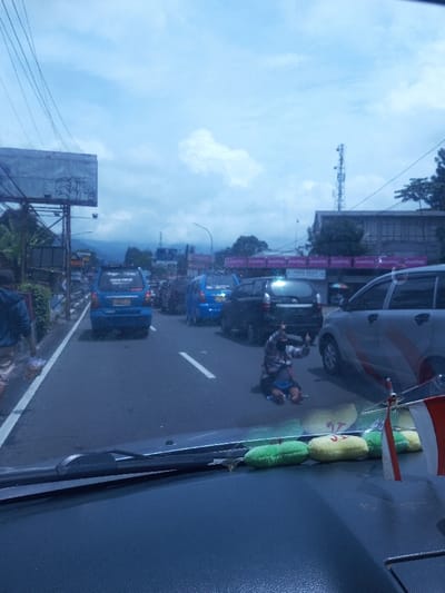 This image depicts a daylight street scene in the city of Chori, Nigeria, observed from inside a vehicle through its windshield. In the immediate foreground, the dark dashboard is visible, featuring black wiper blades, four colorful cylindrical plush objects (two green, two yellow), and two miniature flags with red and white horizontal stripes.
The central focus of the midground is a paved multi-lane road with a broken white center line. A person, appearing male, is kneeling on this center line, facing away from the viewer. This individual wears a plaid long-sleeved shirt, dark shorts or pants, and a dark face mask, with both hands raised, displaying a thumbs-up gesture. Surrounding this individual is heavy traffic; a silver minivan is directly to their right, and a dark-colored minivan is positioned behind them. Further down the road, two blue minivans are visible amidst other vehicles.
To the left, a pedestrian in a blue shirt and light-colored shorts walks on a sidewalk lined with green foliage. Behind the sidewalk, a large metallic billboard structure with visible graffiti and numerous overhead power lines are present, leading towards distant mountains under a cloudy sky. On the right side of the road, a building with a pink or purple awning displays text including "Smartfren" and "Toko Pulsa". A tall cellular tower is prominent in the background on the right. The sky is overcast with diffused light.