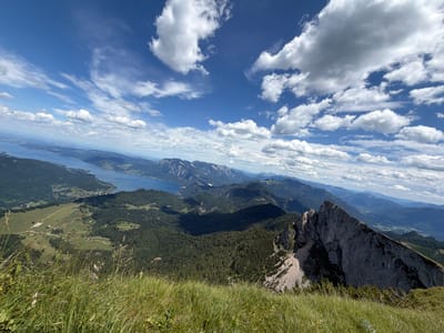 The image depicts a panoramic view from a mountain summit overlooking Sankt Gilgen, Austria. The foreground is dominated by tall, green grass with a few small wildflowers. Beyond the grass, the scene unfolds to reveal a vast mountainous landscape. A large, deep blue lake is visible in the mid-ground, with the town of Sankt Gilgen nestled along its shores. The lake is surrounded by rolling green hills and dense forests, which give way to more rugged, darker mountains in the distance. The sky is a vibrant blue, dotted with fluffy white clouds, suggesting fair weather. A steep, rocky mountain ridge juts out into the foreground on the right side of the image, adding a dramatic element to the otherwise serene vista. The overall impression is one of natural beauty and expansive, untouched wilderness.