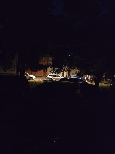 In the city of Kostelec na Hané, Czechia, at night, parked cars are visible on a street in front of buildings. A few people are standing near a black car under a large tree. The scene is dimly lit, with shadows cast by the trees obscuring much of the view. The overall impression is of a quiet street scene at dusk or after dark.