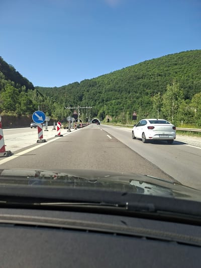 The image captures a view from inside a vehicle, looking forward along a highway. The road is bordered by a steep, densely forested hillside on the left and the edge of the vehicle on the right. The sky is a clear, bright blue, suggesting a sunny day, likely midday.
The scene depicts a stretch of highway with roadwork in progress. Several traffic cones, alternately striped red and white, are set up along the right side of the road, guiding traffic. A blue circular sign with a white arrow pointing straight ahead is prominently visible among the cones, indicating a lane direction. Further ahead, there are traffic lights and what appears to be the entrance to a tunnel.
A white car, possibly a Dacia Logan based on the visible badging, is ahead on the right lane. Its license plate is visible and reads "BG 5730 CK". A reflective barrier runs along the edge of the road. The reflection of the surrounding trees and the sky can be seen on the side of the vehicle in the foreground.
The overall impression is one of a scenic drive through a rural or mountainous area, with temporary road disruptions. The time of day is clearly daytime, and the weather is fair. While no people are directly visible in the frame, the presence of the car and roadwork indicates ongoing human activity. The location context of Vrachesh, Bulgaria, aligns with the visual cues of a highway passing through a forested, potentially mountainous region.