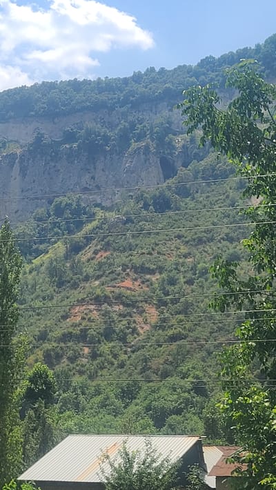 This is a photograph of a mountainous landscape in Arstanbap, Kyrgyzstan. The image captures a verdant hillside dotted with trees and shrubs, with a prominent rocky cliff face in the background. Several utility wires stretch across the frame, indicating human infrastructure within the natural setting. In the foreground, the corrugated metal roof of a building is visible, suggesting residential or agricultural use. The sky above is a clear blue, interspersed with a few wispy clouds. The overall scene depicts a natural environment with signs of human presence.