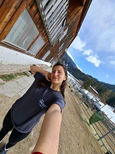 A female individual is depicted taking a selfie outdoors in Davos, Switzerland. She stands centered in the foreground, with dark, wavy hair, a subtle smile, and wears a dark blue t-shirt with light text, black leggings, and dark athletic shoes with white and blue accents. Her left arm is extended forward, holding the camera, while her right arm is raised with her hand near her head. To the left, a multi-story wooden building with dark brown planks, white lower walls, and several windows is visible. To the right, a long metal fence structure with a green net attached extends into the background. The ground is a mix of dirt and dry vegetation. In the background, patches of snow are present on the ground and on the slopes of snow-capped mountains under a blue sky with scattered white clouds and evergreen trees.
