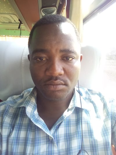 The image shows a man with dark skin, short dark hair, and a mustache, seated inside what appears to be a bus. He is wearing a light blue and white plaid button-up shirt and has earphones in his right ear. His expression is neutral.
Bright sunlight streams in from a window on the right side of the frame, creating a strong highlight on his forehead and causing parts of his face and shirt to be overexposed, indicating it is daytime.
The background reveals the interior of a vehicle, consistent with a bus, featuring light grey seats and an overhead luggage rack with a brown-reddish trim. Inverted green text is visible on the underside of the overhead compartment. While partially obscured and inverted, the text contains characters resembling Swahili words such as "ABIRIA" (passenger), "CHUKUA," and "MAKO," likely forming a notice or instruction for passengers. The overall scene suggests the man is traveling by bus in Sing'isi, Tanzania.