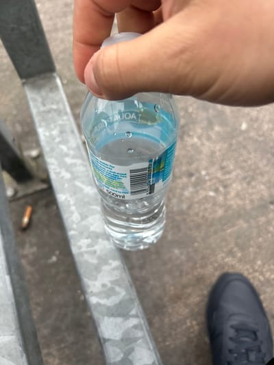 A hand holds a 500ml bottle of "Agua Pura" water over a galvanized metal structure. The bottle's label mentions "brian water for adventures" and its recyclability. A black shoe is visible in the upper right corner of the image, on a concrete surface. A discarded cigarette butt rests near the water bottle on the galvanized metal structure. The scene appears to be outdoors. The location details (Portadown, United Kingdom) are not discernible from the visual information.