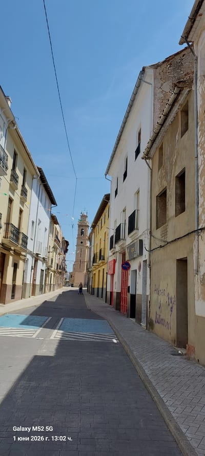A street view in Canals, Spain, shows buildings lining both sides, narrowing towards a central tower. The street pavement is dark brick. Building facades vary in color from white to yellow. A powerline stretches across the sky. A person is walking on the street towards the tower. Graffiti is visible on a building wall. A no parking sign is mounted on a building. The sky is bright blue.
