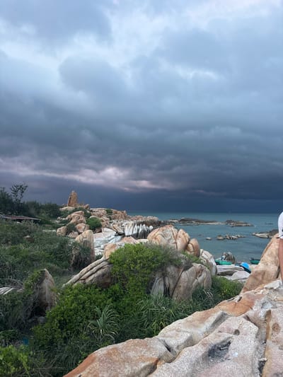 A wide shot shows a rocky coastal landscape with the ocean in the background under a stormy, dark cloud-filled sky. In the foreground, large, light brown, textured rocks are strewn across the landscape, interspersed with lush green vegetation and tall grasses. A few small, green boats are visible in the turquoise water near the shore, which is dotted with more rocks. The sky is dominated by dramatic, dark grey clouds, with a sliver of lighter sky visible on the horizon. A person, partially visible from the waist down and wearing a white shirt, stands on the rocks in the lower right of the frame. The scene appears to be a natural coastal area in Thuận Thành, Vietnam.