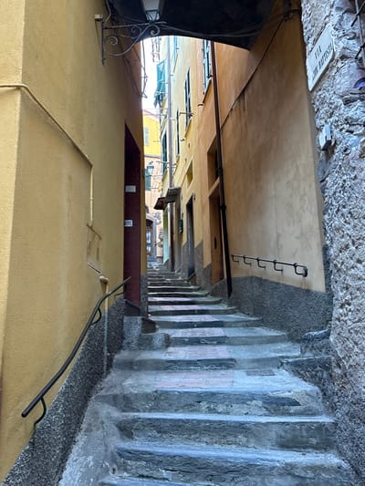 The image depicts a narrow, ascending stone staircase alleyway in Vernazza, Italy, identified by a white street sign on the right reading "VIA G. BATTISTA". The alley is flanked by multi-story buildings painted in shades of yellow and ochre. The immediate foreground features a dark overhead archway, from which a decorative black metal light fixture hangs.
On the left, a smooth yellow stucco wall extends upwards, with a black metal handrail curving along its textured grey base. On the right, the wall transitions from rough, exposed stone in the foreground to a smoother ochre stucco further up. A segmented black metal handrail is affixed horizontally to this wall section.
Approximately 15-20 irregular stone steps ascend into the background, some appearing to have lighter-colored, possibly brick or concrete, inserts. Several dark doorways or entrances are visible along the walls. Further up the alley, windows with green louvred shutters are discernible on the upper floors of buildings. Overhead wires are visible along the building facades. The alley gradually narrows and brightens towards the distant top, where a clear sky is visible. No individuals, animals, or vehicles are present in the scene.