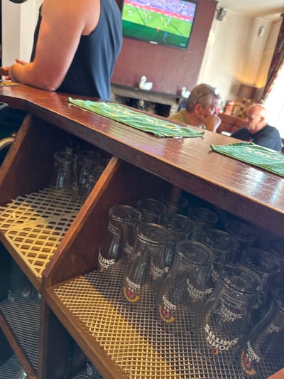 The image depicts the interior of a public house located in Sigglesthorne, United Kingdom. The immediate foreground features a polished wooden bar counter, angled from lower left to upper right, with two green patterned bar towels placed upon it. Below the counter, a wooden shelving unit contains multiple empty glass tumblers and pint glasses, predominantly displaying the "JOHN SMITH'S SMOOTH" brand, resting on white mesh mats.

On the left side of the bar, a person wearing a dark blue sleeveless top is partially visible, with their forearms resting on the counter. In the background, a flat-screen television mounted on a reddish-brown wall displays a football match, with a "0-0" score visible on the screen. Beneath the television, a mantlepiece or shelf holds two white ceramic swan figurines.

Further into the background, two adult males are seated at tables, out of focus. The man on the left has light-colored short hair and glasses on his head, with his hand near his chin. He faces a bald man in a dark short-sleeved shirt seated opposite him, both appearing to be engaged in conversation. A patterned curtain and a lamp are visible on the far right background.