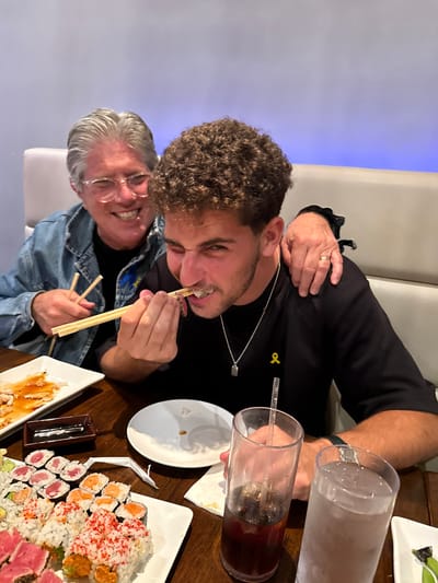An indoor photograph, taken in a restaurant in West Windsor, United States, depicts two individuals seated at a wooden table during a meal. In the foreground, a younger adult male with curly brown hair, wearing a black t-shirt with a yellow ribbon pin and a silver necklace, consumes food. He holds a piece of sushi with wooden chopsticks to his open mouth, displaying a squinted expression and visible orthodontic brackets on his upper teeth. His left arm is bent, holding the chopsticks, while his right arm is largely obscured. To his left, partially behind him, an older adult with silver hair and glasses wears a blue denim jacket over a dark shirt, also featuring a yellow ribbon pin. This individual smiles broadly, holding a pair of chopsticks in their left hand, with their right hand resting on the younger adult's left shoulder.
The wooden table is laden with multiple white rectangular plates showcasing various sushi rolls, including tuna, salmon, and red roe toppings. Another rectangular plate contains a sauced protein dish. A small black dish holds dark liquid (soy sauce). Two clear plastic tumblers containing beverages are present: one with a dark carbonated drink and a straw, and another with a lighter, possibly iced, liquid. An empty white plate and crumpled white napkins are also visible. The background consists of light-colored booth seating and a wall illuminated with a soft blue ambient light.