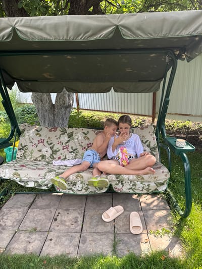 Two children, a boy and a girl, sit on a floral-patterned swing seat in an outdoor setting. The boy, wearing blue shorts, is on the left, and the girl, wearing a white top and patterned shorts, is on the right. They are both looking down at a bag of chips, with the girl holding it and offering some to the boy, who is reaching for them. The swing is green and has a canopy overhead. In front of the swing, on a paved area, are two beige slides. The background shows greenery, trees, and a white fence. The image appears to be taken in the village of Nizhne-Chufichevo, Russia, during daylight hours.