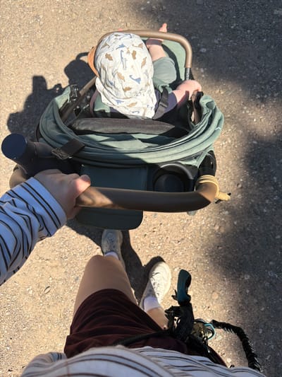 A first-person perspective shot shows a person pushing a stroller on a gravel path. The stroller contains a baby wearing a dinosaur-patterned hat and green pants. The person pushing the stroller is wearing a striped long-sleeved shirt, maroon shorts, and white sneakers. A rope is attached to the stroller handle. The scene appears to be outdoors in Odernheim am Glan, Germany, with sunlight casting shadows on the ground.