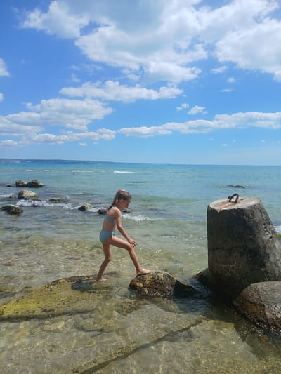 A young girl in a blue bikini stands in shallow, clear water near the shore. She is positioned on a rock, with her right leg extended and her left leg bent at the knee, resting on another rock. Her arms are bent and held slightly away from her body. She is looking towards the water. The scene is a beach with a clear blue sky filled with fluffy white clouds. The sea is calm with gentle waves lapping the shore. Several rocks are visible in the water and along the shoreline. To the right of the frame, a large, weathered concrete structure, possibly a breakwater or part of a pier, is visible. The sun appears to be high in the sky, suggesting it is daytime. The overall mood appears peaceful and summery. The location is Osenovo, Bulgaria. There is no visible text.