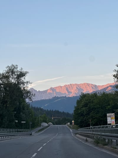 The image depicts a scenic view of a road leading towards a mountain range, likely in or near Farchant, Germany, given the presence of German signage. The sun is setting, casting a warm, pink glow on the rugged mountain peaks in the distance. Lush green trees line both sides of the road, with one side featuring a dense forest and the other a slightly sparser arrangement. The road itself is asphalt, with clear white lane markings, and appears to be an elevated section, possibly a bridge, judging by the metal railings on either side. Several streetlights are visible along the roadside. A signpost with multiple directional signs in German is also present on the right side of the road. The sky is a clear, pale blue, with a faint streak of a contrail visible in the upper portion. The overall atmosphere is peaceful and serene, capturing a moment of natural beauty enhanced by the golden hour light.