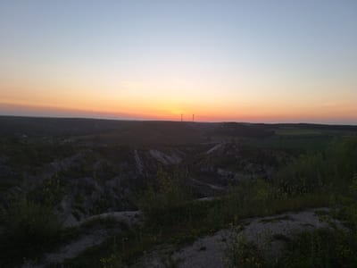 This image captures a tranquil sunset scene in Fetești, Moldova. The foreground features a rocky, overgrown path with sparse vegetation, suggesting a natural or slightly wild landscape. Beyond this, the terrain dramatically slopes downwards into a valley, characterized by eroded, lighter-colored slopes dotted with greenery. In the mid-ground, a winding road snakes through the undulating hills.
Dominating the horizon are two prominent wind turbines, silhouetted against the vibrant sunset sky. The sky itself transitions from a pale blue at the top to a warm gradient of orange and pink hues near the horizon, indicative of the twilight hour. The overall impression is one of peacefulness and natural beauty, with the wind turbines serving as a modern element within the rustic setting. The time of day is clearly sunset, and the weather appears clear and calm. There is no visible text in the image.