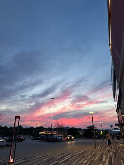 Captured from a ground-level perspective, the image portrays an urban evening scene in Oak Brook, Illinois, USA. The sky features vibrant hues of pink and grey clouds, indicating either a sunset or sunrise. On the right side, a paved walkway with a geometric pattern leads to a street lined with parked cars and mature trees. Street lamps illuminate the walkway and street. People are walking on the walkway in the distance, and a water tower is visible. A building is visible on the left side of the image.