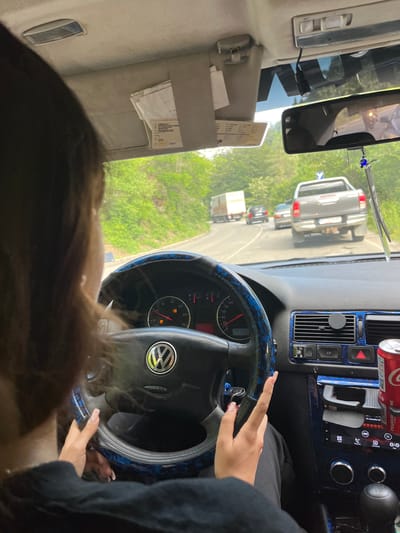 The image shows the interior of a Volkswagen car, viewed from the passenger's perspective. A person, only partially visible, is driving. The dashboard features a steering wheel with a Volkswagen logo, a speedometer, and various controls. Decorative blue trim is visible on the steering wheel and dashboard. Outside the car, a road is visible with other vehicles, including a truck and a pickup truck, and trees lining the roadside, suggesting a rural or suburban setting. A can of Coca-Cola is placed in a cup holder. The location is identified as Vlahovo, Bulgaria.