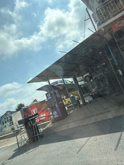 This media file depicts a vibrant street scene in Vresovo, Bulgaria, captured at a tilted angle through a reflective surface, likely a vehicle window. The sky, filled with scattered white clouds, is visible in the upper left portion of the image, with bright sunlight indicating daytime. Reflections of the sky and clouds are evident across the entire frame.

On the right side of the image, a paved street or sidewalk runs along what appears to be a series of storefronts or businesses. A person is dimly visible in the background, near one of the establishments. Several signs and objects are prominent. There are multiple blue-toned signs advertising "LAVAZZA" coffee, suggesting a coffee shop or a location selling Lavazza products. Below these, a large red banner features a stylized "F" logo and the text "IZJID DEALER," possibly indicating an official dealer for a car brand or automotive parts.

Further along, a sign listing various services is visible. The OCR identifies these as: "ТЕЛ.: 0884 53 53 22" (a phone number), "2 СМЯНА НА МАСЛА" (Oil Change), "3. ЗАВАРКИ НА АУСПУСИ" (Muffler Welding), and "4 СТИЛЯРНА СИСТЕМА" (Styling System). This suggests the presence of an auto service or repair shop. An upside-down metal structure, possibly a chair or a display rack, is on the sidewalk near the auto service sign. The general impression is one of a commercial street with a mix of businesses, including automotive services and a coffee outlet, under a clear, bright sky.
