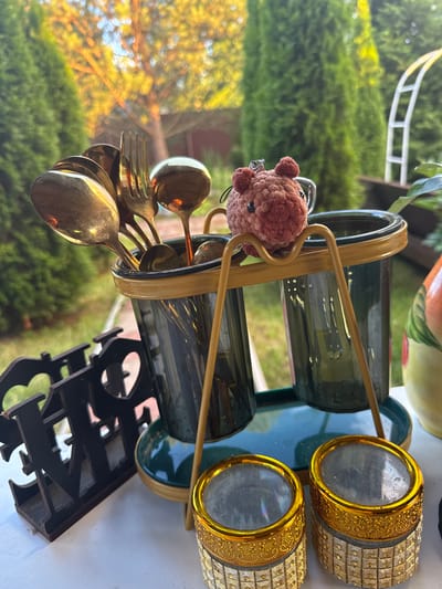 The image shows a close-up of a cutlery holder filled with various metallic spoons and a fork, all with a golden sheen. A small, crocheted brown teddy bear charm is attached to the top of the holder by a metal ring. The holder itself is gold-colored metal with dark green glass inserts, resting on a green platter. To the right of the holder are two decorative gold and silver containers, possibly for storing items. In the background, lush green trees and foliage are visible, suggesting an outdoor setting. Sunlight filters through the leaves, creating a warm, dappled effect. A portion of a white fence or railing can be seen in the upper left corner, and a black, ornate decorative item is visible in the lower right. The overall impression is one of a decorative outdoor dining or picnic setup. There is no visible text.