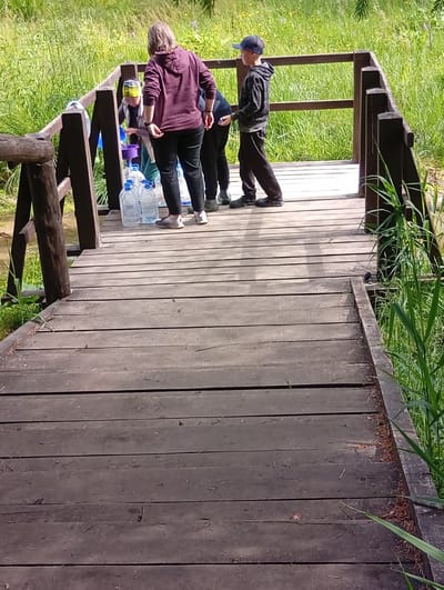 In Svilė, Lithuania, a group of people stand on a wooden bridge, possibly collecting water from a nearby source. The scene is set amidst lush green foliage and tall grass, suggesting a natural, outdoor environment. The bridge is constructed of weathered wooden planks, with a sturdy wooden railing.
Three individuals are visible. One person, wearing a maroon hoodie and black pants, stands on the bridge with their back to the camera, attending to several large plastic water bottles. Another person, in a dark hoodie and black pants, is also present, wearing a dark baseball cap. A third person, partially obscured, is seen in a blue shirt and a yellow bandana.
Several large, clear plastic water bottles are lined up on the bridge, some of which appear to be full or being filled. A purple bucket is also visible near the person in the maroon hoodie, suggesting a water collection or purification activity. The sunlight filtering through the trees and the vibrant green of the surrounding nature indicate it is daytime, likely a warm and pleasant day. The atmosphere seems focused and practical, with no overt emotional displays. The wooden structure of the bridge and the natural surroundings hint at a rural or park-like setting. There is no visible text in the image.