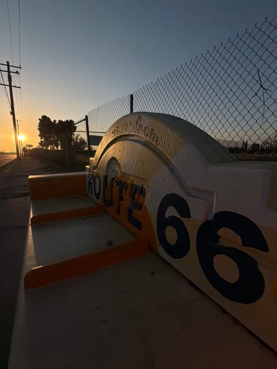 The image displays an outdoor scene in Fontana, United States of America, captured during either sunrise or sunset. In the immediate foreground, a weathered, low-lying monument or bench is visible, constructed with a cream-yellow base, orange trim, and a prominent dark blue/black "ROUTE 66" inscription, with "ROUTE" partially obscured on a curved section and "66" clearly visible on the right. The object is illuminated by direct, low-angle sunlight, creating bright highlights and casting partial shadows.

Behind the monument, a chain-link fence with pointed security elements extends across the midground, separating the foreground from distant features. To the left, a utility pole with multiple overhead wires stands silhouetted against a brilliant, low sun visible on the horizon. Sparse, dark trees are observed behind the fence, with faint, hazy hills or mountains discernible in the far background. The sky transitions from a vibrant orange-yellow at the horizon to a pale blue overhead. No individuals or active subjects are present in the scene.
