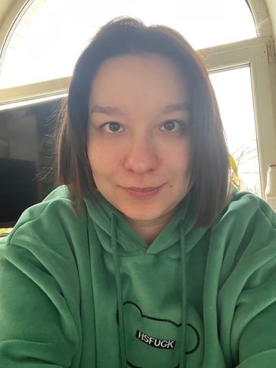 An adult female individual is centered in a close-up indoor photograph, likely a selfie, taken in Kozlovka, Russia. She has short, dark brown hair parted on the side, light brown eyes, and a neutral expression with a slight smile. She is wearing a dark green hooded sweatshirt, or hoodie, made of a textured, possibly velour material, with visible drawstrings. The hoodie features an embroidered design on the front depicting a stylized bear outline containing the text "HSFUCK" in white capital letters on a black patch. The background on the left consists of a dark, reflective surface, possibly a television screen. The background on the right features a white-framed window admitting bright daylight, with a plant in a pot and a red-capped bottle partially visible near the window frame. The overall lighting is bright, with backlighting from the window.