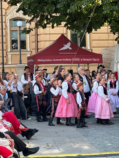 A group of children and adults are participating in a cultural event, possibly a festival or performance, in front of a building in the city of Nowy Sącz, Poland. Many of the participants, particularly the children, are dressed in traditional folk costumes. These costumes include embroidered vests, white shirts, and dark pants or skirts, with some boys wearing straw hats. Several of the children are wearing bright pink skirts. Adults in the background are also dressed in similar traditional attire or more casual clothing. A red tent with text indicating "Małopolskie Centrum Kultury SOKÓŁ w Nowym Sączu" (Malopolska Cultural Center SOKOL in Nowy Sacz) is set up behind the performers. The event is taking place outdoors on a paved area, with trees and a building visible in the background. Some spectators are seated in the foreground, observing the event, while others are standing and taking photos. The overall scene suggests a lively cultural gathering in a public square in Nowy Sącz.