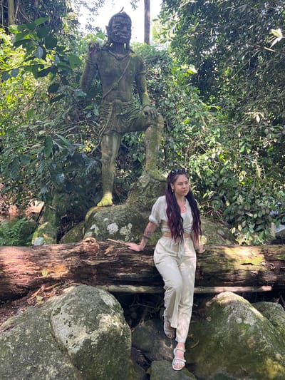 A woman in a light-colored outfit with a tied-front top and loose pants is sitting on a fallen log in front of a moss-covered stone statue of a warrior. The statue is in a jungle environment with lush green foliage and dappled sunlight. Large rocks are scattered around the base of the statue and the log. The woman has long, dark hair with reddish highlights and is wearing sandals. Her left arm rests on the log, and her right hand is also near the log. The statue appears to be ancient and weathered. The scene is set in the city of Ban Bang Khao, Thailand.