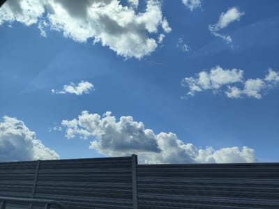 The image shows a clear blue sky filled with white and gray clouds, with a distant contrail from an airplane visible. In the foreground, a dark gray sound barrier fence runs horizontally across the frame. The fence has a ribbed texture and is supported by vertical concrete pillars. A metal railing is visible at the bottom left corner. The scene appears to be taken from a vehicle, as indicated by a slight reflection on the sky. The location is identified as Katlakalns, Latvia.