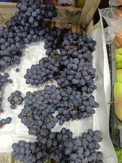 The image shows a top-down view of a large pile of dark purple grapes. The grapes are arranged in several bunches, some of which are quite large and dense, while others are smaller and more scattered. The grapes are resting on a white surface, which appears to be paper or cloth. In the background, a wooden crate is visible, and behind that, some plastic packaging and other produce, possibly oranges and pears, are present. The lighting suggests an indoor market or grocery store setting. The location is specified as a market in Gulbakhor, Uzbekistan. There are no people visible in the image.