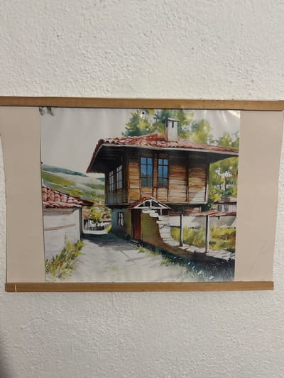This is a watercolor painting of a village scene in Shipkovo, Bulgaria. The painting depicts a narrow, paved pathway winding between two traditional houses. The house on the right is constructed of wood with a red-tiled roof and features prominent windows with blue panes. A white chimney rises from its roof. The house on the left is white-walled with a red-tiled roof and a white stripe along its facade. Greenery, including trees and bushes, is visible around the houses and in the background, where rolling hills are depicted under a bright sky. The overall impression is one of a sunny, rural setting. The painting is framed by a thin wooden border.