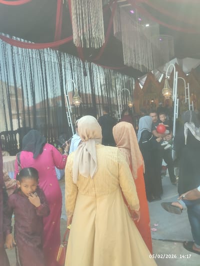 A group of people are gathered at what appears to be an event or celebration. Several women are wearing traditional dresses and headscarves, with one child in a purple dress. A man is holding a toddler with a red balloon. The scene is decorated with curtains and hanging ornaments. A date stamp in the corner of the image reads "05/02/2026".