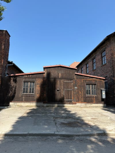 This is an outdoor, eye-level, medium shot of a rustic wooden building with a brick building partially visible to the left, set against a clear blue sky. The wooden structure has several windows with metal bars, suggesting security or confinement. Its dark wood is weathered and shows vertical planking. The brick building is older, with visible rows of bricks and multiple windows. The scene is illuminated by bright sunlight, casting strong shadows across the concrete ground on the right, indicating a sunny day. The overall impression is one of historical significance, possibly a former prison or industrial site. There are no people visible in the frame. The location context of Oświęcim, Poland, suggests this could be a building within the Auschwitz-Birkenau Memorial and Museum. No text is visible in the image.