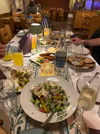 The image depicts an interior dining scene at Restaurant Poseidon, a Greek restaurant located in Eppelheim, Germany. The primary focus is a dining table covered with a white tablecloth and an olive branch-patterned runner, illuminated by warm artificial light, including a green portable table lamp and a lit candle in a glass holder.

The table is laden with various dishes and beverages. Prominently displayed is a large white plate containing a Greek salad composed of cucumber, tomato, red onion, feta cheese, olives, and green peppers. Adjacent to it are a plate of grilled green chili peppers (peperoni) with a white sauce, and another plate holding three fried, breaded items garnished with a lemon wedge and small salad. A plate of toasted bread slices is also visible. Beverages include a bottle of "SELTERS Classic" sparkling water, two glasses of orange juice, and multiple champagne flutes, some containing a light yellow liquid consistent with white or sparkling wine. A glass with a "Havana Club" logo is also present. A small Greek flag stands upright on the table.

In the left foreground, a human hand is holding a fork, actively engaging with the plate of grilled peppers. In the midground, other blurred plates of food and glasses are discernible. The background shows the blurred interior of the restaurant, populated by other diners seated at wooden tables and chairs, and a standing individual in a plaid shirt further in the distance. The overall environment suggests an evening dining service.