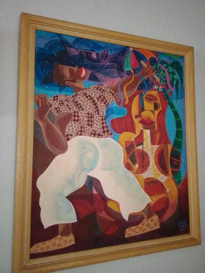 The image displays a vibrant, colorful painting enclosed in a light wooden frame, hanging on a plain, light-colored wall. The painting depicts a stylized male figure actively dancing or posing in a celebratory manner, characteristic of a Carnival or festive scene.

The central figure, with dark skin, wears a patterned shirt featuring both polka dots and diamond shapes in muted tones, and loose-fitting white pants. His shoes are also patterned with polka dots. He has a wide, joyful smile with bright red lips and prominent white teeth. His right arm is bent, seemingly holding a small, multi-colored parasol, while his left arm extends forward with colorful ribbons or streamers flowing from it.

The background is a dynamic composition of geometric shapes and colors. The upper left features a dark blue area speckled with white dots, suggesting a night sky with stars or confetti. To the right of the central figure, a more abstract, anthropomorphic shape in shades of red, yellow, and orange with distinct red lips and dark eyes, possibly representing another figure or a musical instrument, is visible. A green palm tree or tropical plant stands on the far right. The overall color palette is rich and lively, dominated by blues, reds, yellows, and greens.

In the bottom right corner of the painting, a signature reads "ALMIRA RAMOS 17", indicating the artist and potentially the year 2017. The physical setting of the painting itself is a plain, light-colored wall. No specific environmental cues within the photograph indicate its precise location in the city of Campo Grande, Brazil, beyond the assumption provided in the prompt.