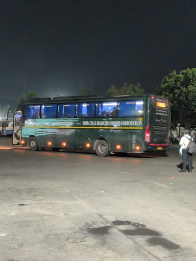 The image shows a green Himachal Road Transport Corporation (HRTC) bus parked on a paved surface, likely a bus depot or parking lot, at night. The bus is illuminated by its internal lights and external hazard lights, with trees and a dark sky in the background. Two individuals are visible. One person, wearing a light-colored shirt and dark pants with a backpack, stands facing away from the camera. Another person, wearing a dark uniform, is visible near the open door of the bus, appearing to be an attendant or driver. The bus has "HIMACHAL ROAD TRANSPORT CORPORATION" written in white on its side. The rear of the bus displays "HRTC" and a route destination in amber illuminated text. There are puddles of water and debris on the ground. The scene appears to be in Kajheri, India, a location known for its bus station.