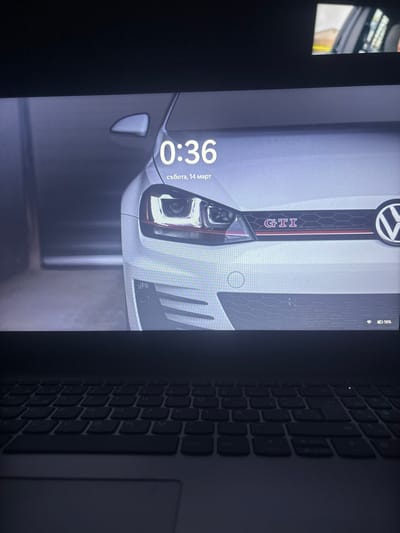 The image displays a photograph of a laptop computer screen and the upper portion of its keyboard and touchpad. The screen exhibits a digital lock screen featuring the front fascia of a white Volkswagen Golf GTI. Visible elements of the car include the left headlight assembly, part of the front grille with a red horizontal accent stripe and "GTI" emblem, and the Volkswagen logo on the right side. Overlaid on this background, the time "0:36" is displayed prominently, with "събота, 14 март" (Saturday, March 14) in Bulgarian below it. In the bottom right corner of the screen, a battery icon indicates a 58% charge, alongside a Wi-Fi signal icon. The laptop's dark-colored QWERTY keyboard and a portion of the touchpad are visible in the lower section of the photograph, appearing dimly lit. In the top right corner of the overall image, a partial, blurred view of an interior car door frame, possibly a reflection, is present. No individuals are depicted. The image itself contains no visual indicators identifying the physical location as Tsenovo, Bulgaria.